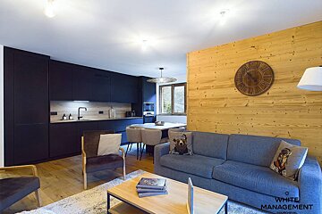 Modern open-plan living room and kitchen with dark blue cabinets, light wood walls, grey sofa, and dining area, featuring a cozy chalet style.