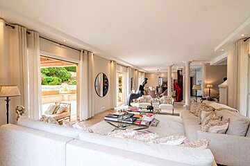 A spacious, elegant living room in light beige tones, featuring modern decor and open glass doors leading to a patio.
