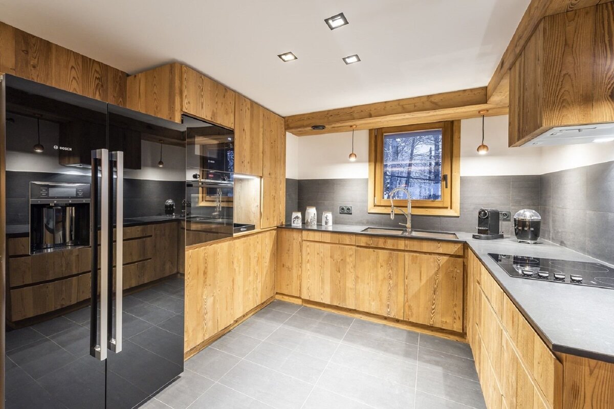 A modern kitchen features light wooden cabinets, dark gray counters & floor, sleek black appliances, and a window overlooking a snowy landscape.