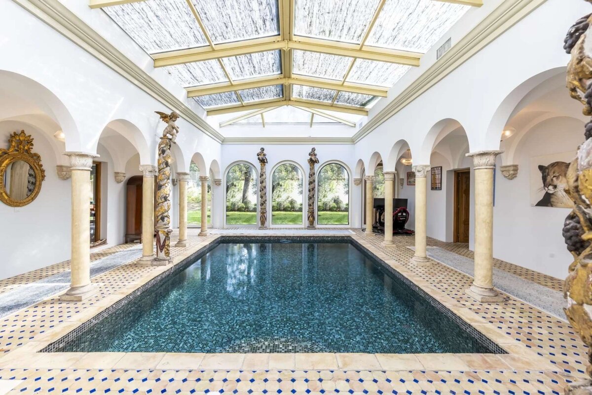 An elegant indoor pool with arched columns, a skylight roof, and ornate details, looking out to a garden through large windows.
