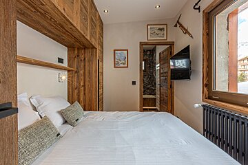 Chalet-style bedroom with a white bed, wood paneling, wall TV, window, and an open door revealing a bathroom.