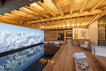 A warm, rustic indoor pool area in a wooden chalet. A large mountain mural reflects in the water, with loungers nearby for relaxation.