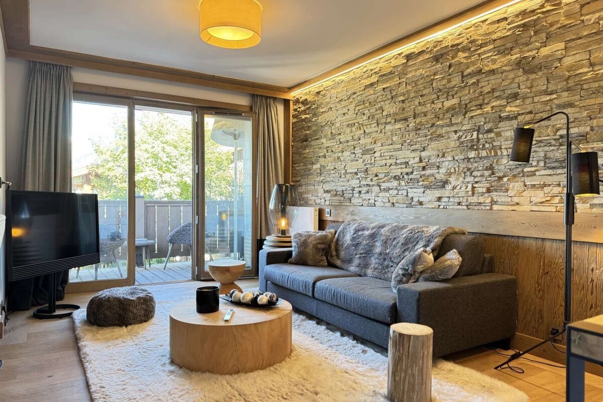 A cozy living room featuring a stone accent wall, grey sofa with fur throws, round wooden coffee table, plush rug, and a balcony view.
