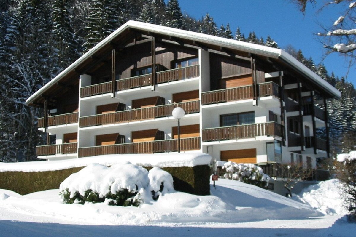 A large, modern chalet with balconies, covered in snow, nestled among snow-covered pine trees under a clear blue sky in winter.