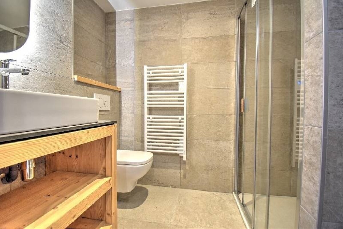 A modern bathroom with grey tiles, a wooden vanity with a white sink, a toilet, a heated towel rack, and a glass-enclosed shower.