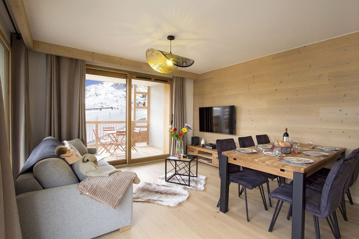 A cozy, modern living/dining room with light wood walls & floors. Features a grey sofa, dining table, TV, and a balcony with snow mountain views.