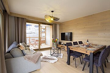 A cozy, modern living/dining room with light wood walls & floors. Features a grey sofa, dining table, TV, and a balcony with snow mountain views.