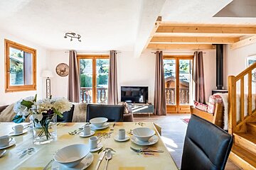 Bright, cozy chalet interior featuring a set dining table with white bowls, leading into a sunny living room with glass balcony doors.