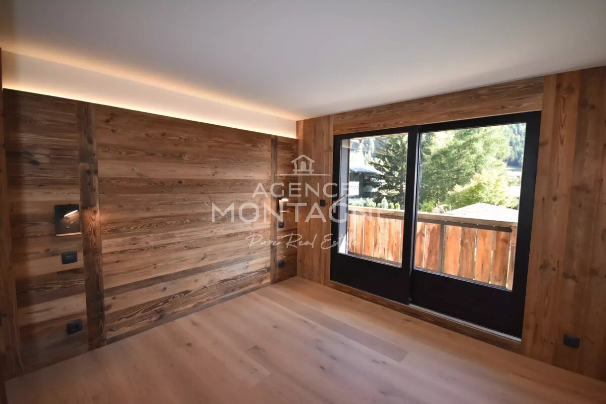 A rustic room with wood-paneled walls, light wood floor, and large sliding glass doors opening to a balcony with a green view. Subtle ceiling lighting.