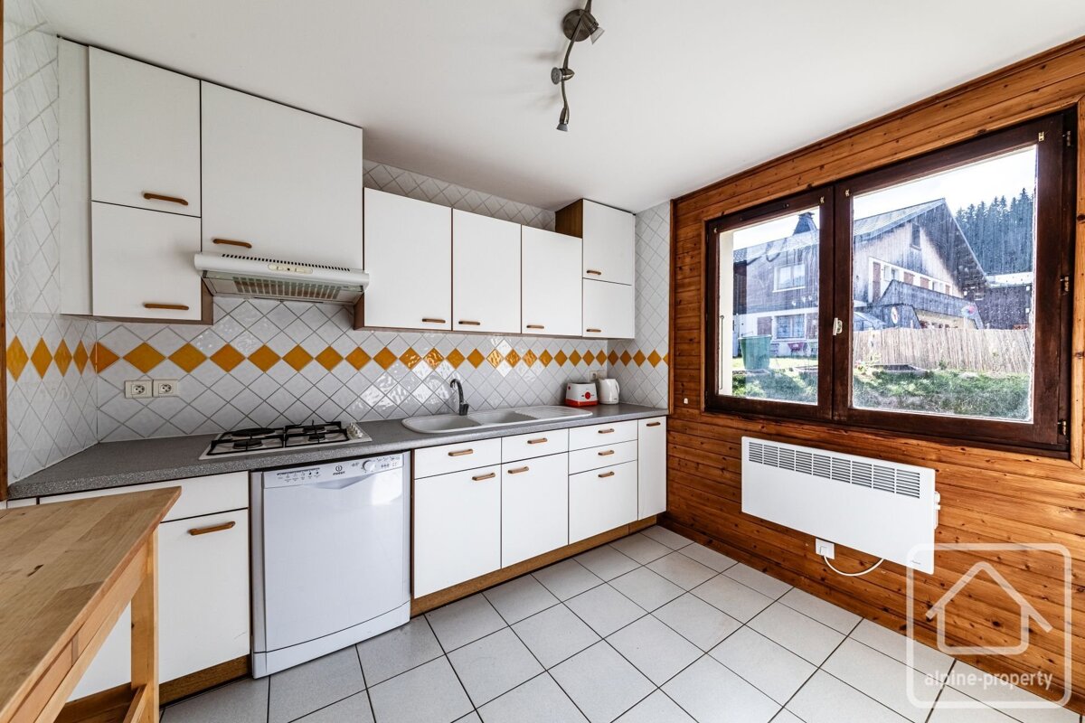 A bright kitchen with white cabinets, a yellow and white diamond-patterned backsplash, and a rustic wooden wall with a large window. It has a tiled floor.