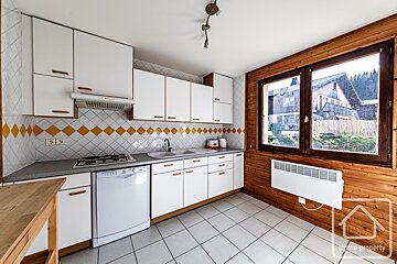 A bright kitchen with white cabinets, a yellow and white diamond-patterned backsplash, and a rustic wooden wall with a large window. It has a tiled floor.