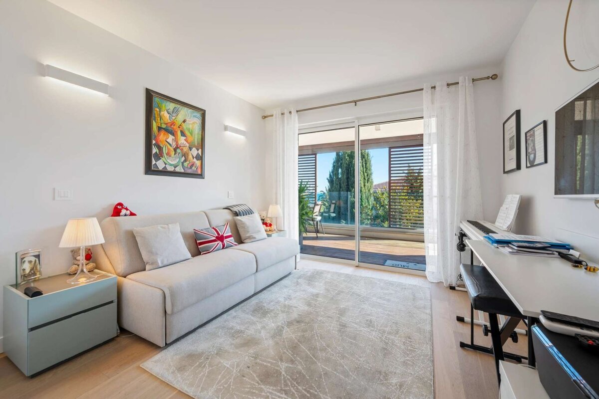 Bright living room with a light gray sofa, desk with a keyboard, and large glass doors opening to a balcony with trees and a blue sky.