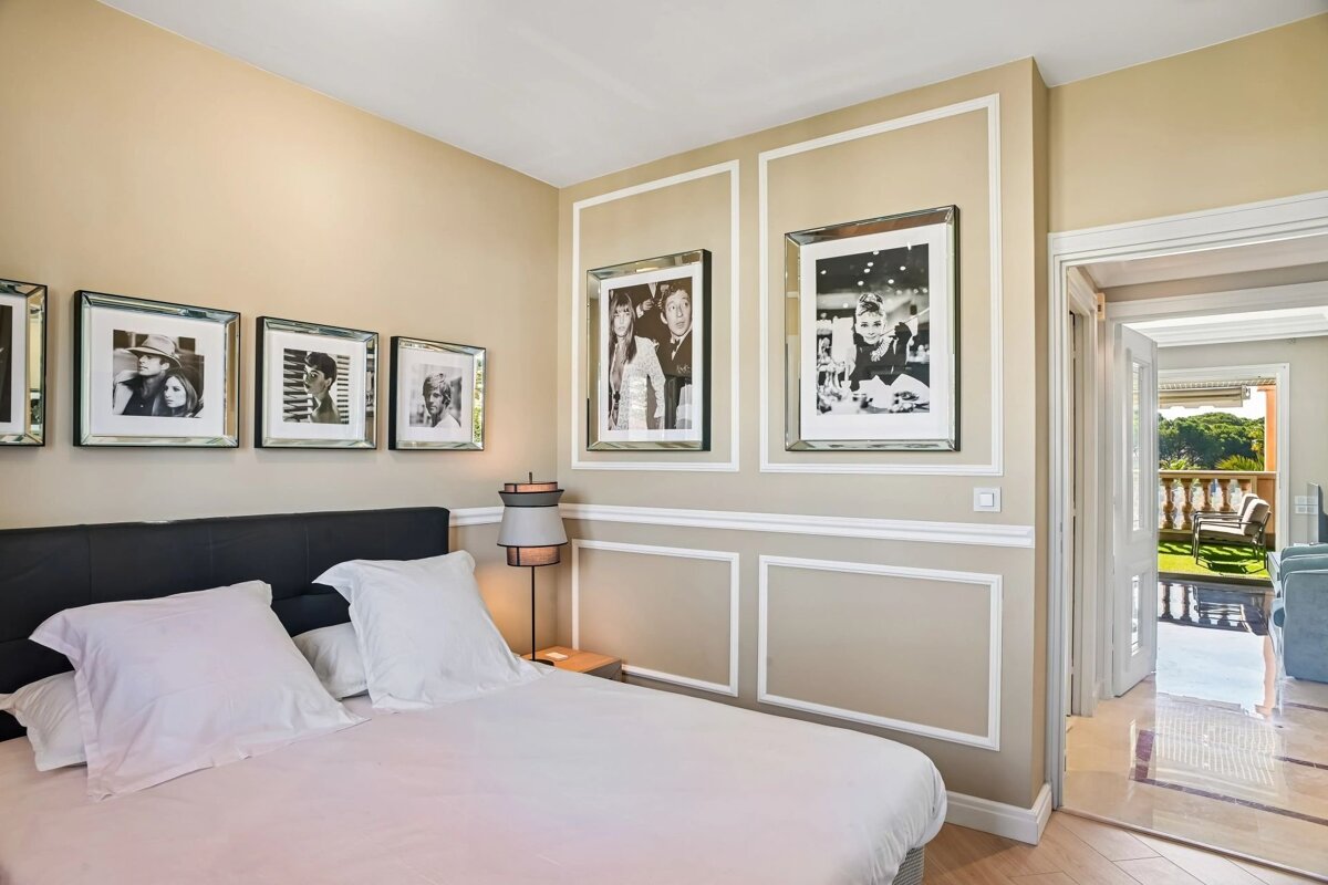 An elegant bedroom with beige walls, black and white framed portraits, a black headboard, and an open doorway leading to a sunny balcony.