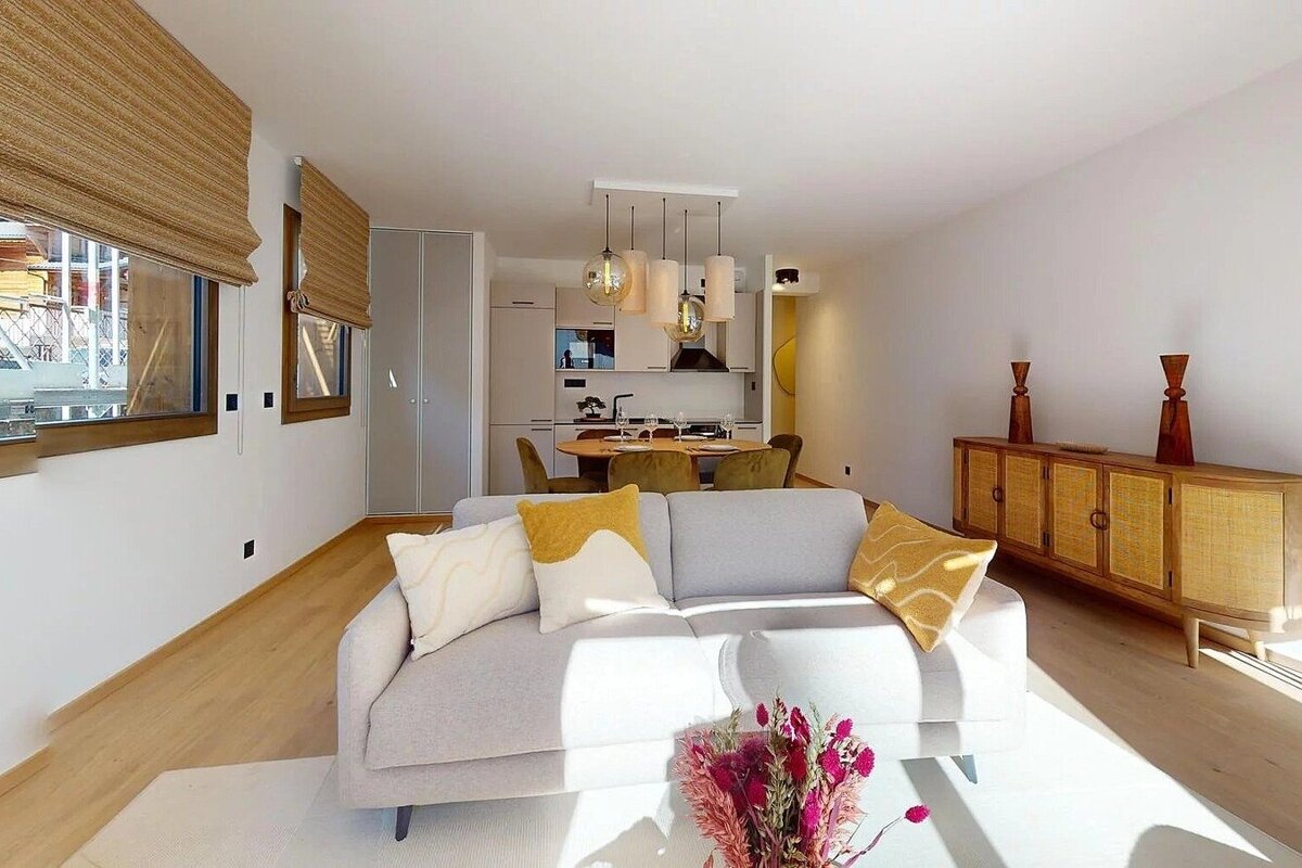 Bright, open-plan living/dining/kitchen with a light gray sofa, yellow cushions, and wood dining set. A modern kitchen and wood/rattan sideboard complete the contemporary, natural-toned space.