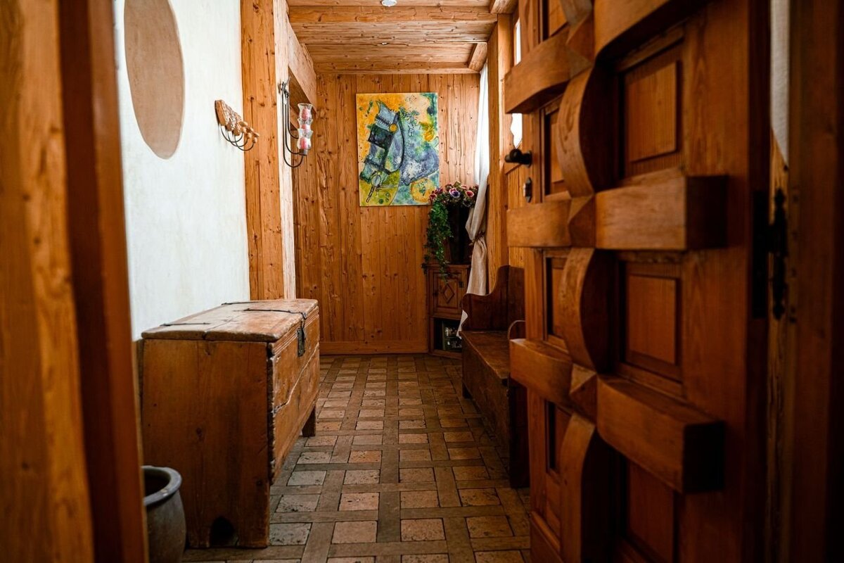 A rustic hallway with wooden walls, ceiling, and an ornate door. It features a tiled floor, a large wooden chest, a bench, and a colorful painting.