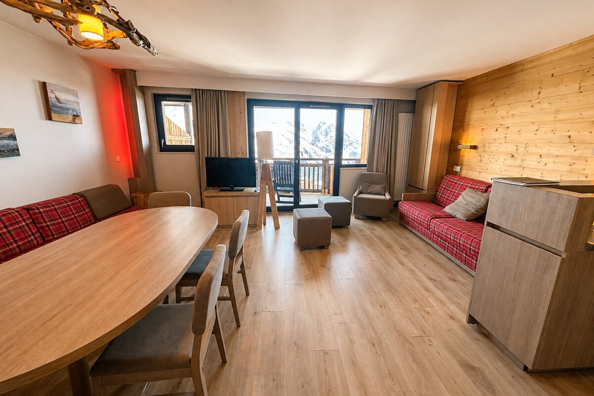 A rustic chalet living area with wood floors, a dining table, a red plaid sofa, and views of snowy mountains.