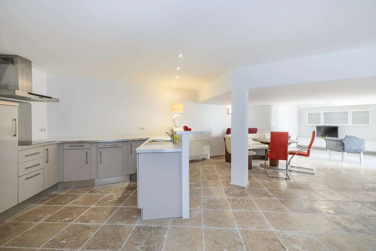 An open-plan living space with a modern kitchen featuring grey cabinets, a dining area with red chairs, and a lounge, all on a tiled floor.