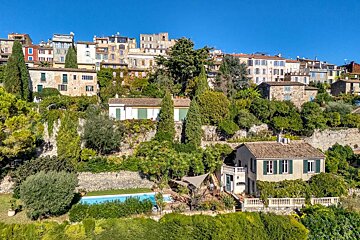 A picturesque village built into a lush hillside, with traditional houses, dense greenery, and a blue swimming pool in the foreground.