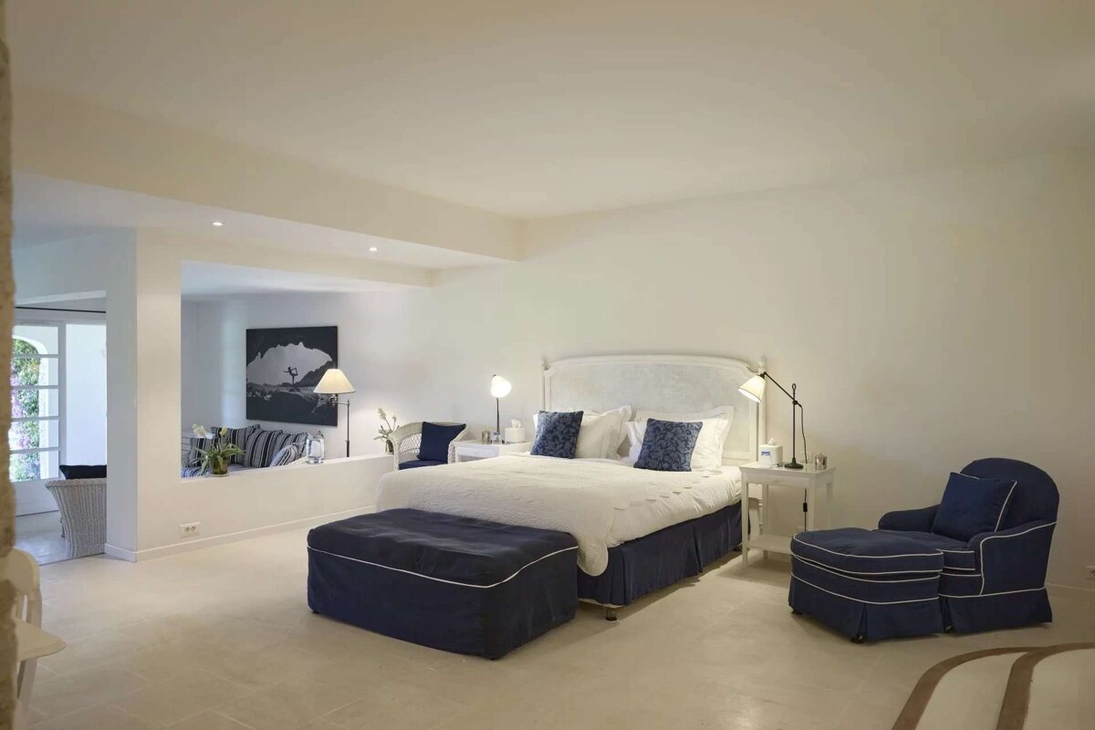 A bright, modern bedroom featuring a white bed with blue accents, an ottoman, and an armchair, with an open sitting area beyond a low wall.