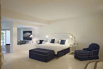 A bright, modern bedroom featuring a white bed with blue accents, an ottoman, and an armchair, with an open sitting area beyond a low wall.