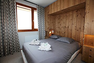 A cozy bedroom features wooden paneling above a grey bed with decorative towels, flanked by patterned curtains and a bright window.
