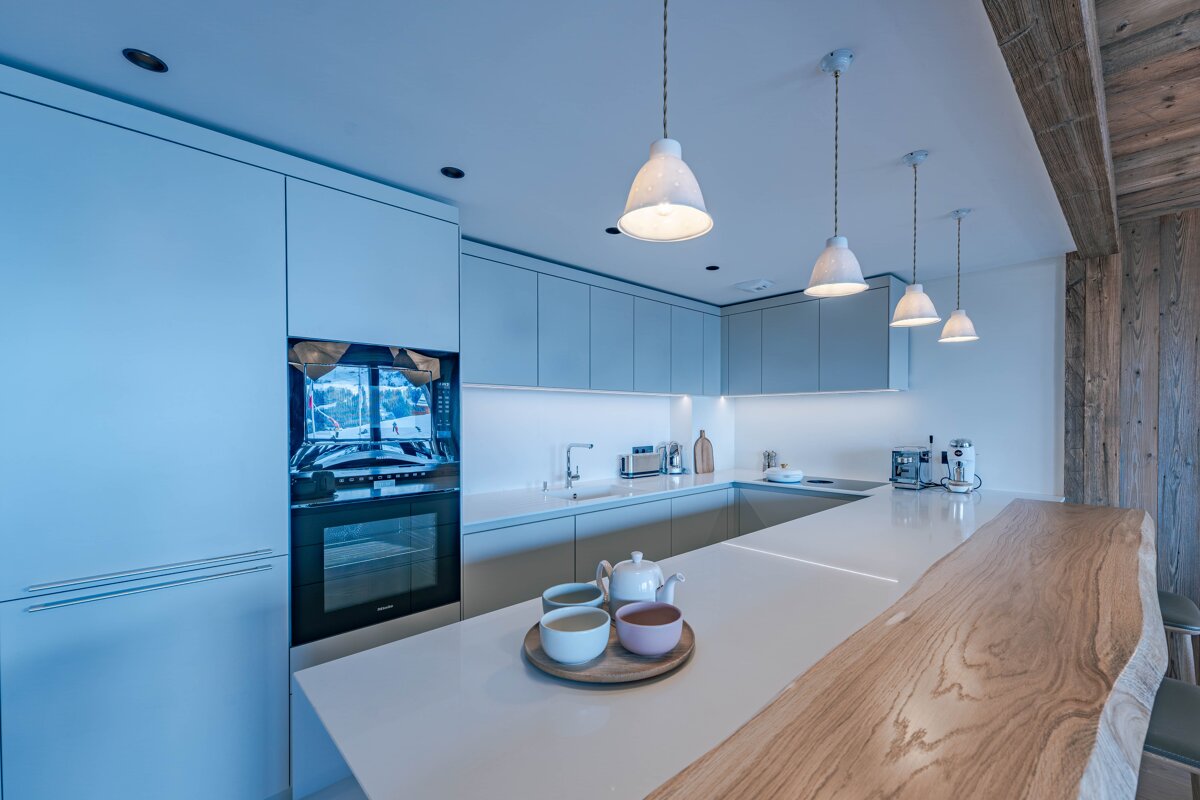 A sleek, modern kitchen featuring pale blue-grey cabinets, white countertops, stylish pendant lights, and a natural wood bar. Teapot and cups sit on the island.