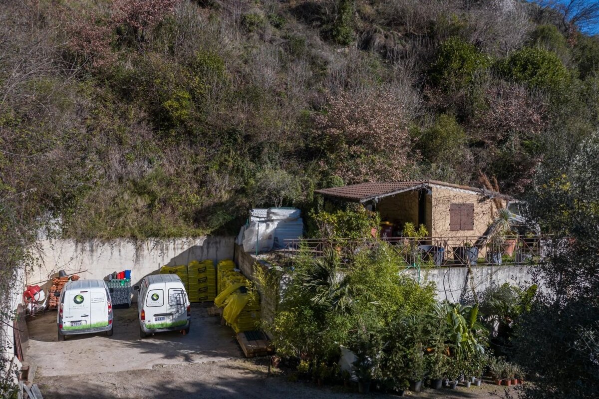 A rustic house with a tiled roof is nestled against a steep, overgrown hill. Two white vans, gardening supplies, and many potted plants are visible.