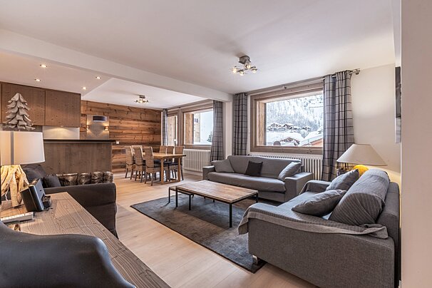 Cozy chalet living room with grey sofas, a wooden dining set, and large windows looking out onto a snowy mountain landscape.