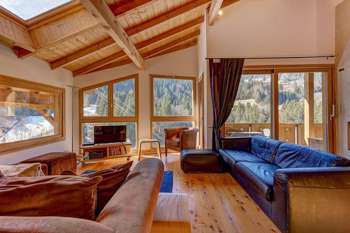 A cozy, rustic living room with wooden beams, large windows offering scenic mountain and forest views, and comfortable seating.