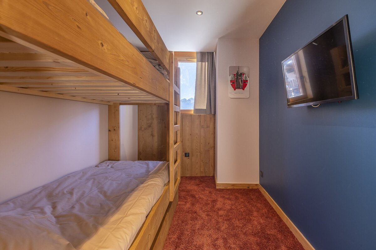 A small bedroom featuring wooden bunk beds, red carpet, a blue wall with a mounted TV, and a window at the far end.