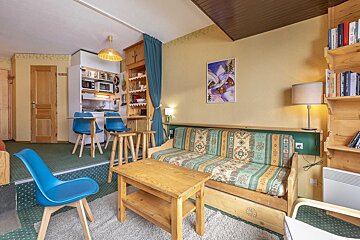 A cozy, rustic living room with a patterned sofa, wooden furniture, and a compact kitchen area. Features yellow walls, green carpet, and ski-themed art.