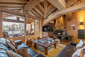 Cozy alpine chalet living room with exposed wooden beams, leather sofas, a warm fireplace, and winter views through large windows.