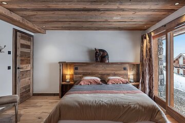A cozy rustic bedroom with wood-paneled ceilings, a wooden bed, a horse head sculpture, and sliding glass doors with a snowy view.