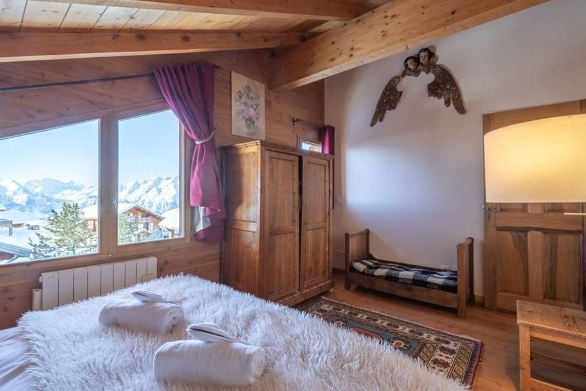 Cozy wooden alpine bedroom with snowy mountain views, a fluffy bed with rolled towels, rustic wardrobe, and warm lighting.