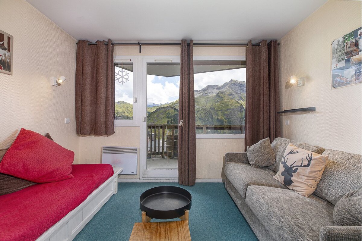Cozy mountain apartment with a red futon, grey sofa, blue carpet, and a glass door opening to stunning, rugged mountain views.