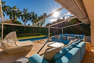 A bright, sunny patio with blue couches, a white daybed, and a swimming pool framed by palm trees.