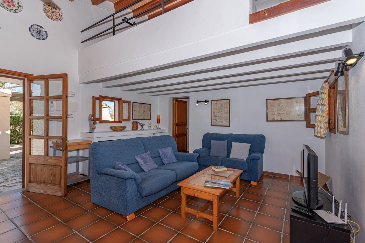 A rustic living room with blue couches, terracotta floor, and wood-beamed ceiling. Features a wooden coffee table, TV, and open doorway.