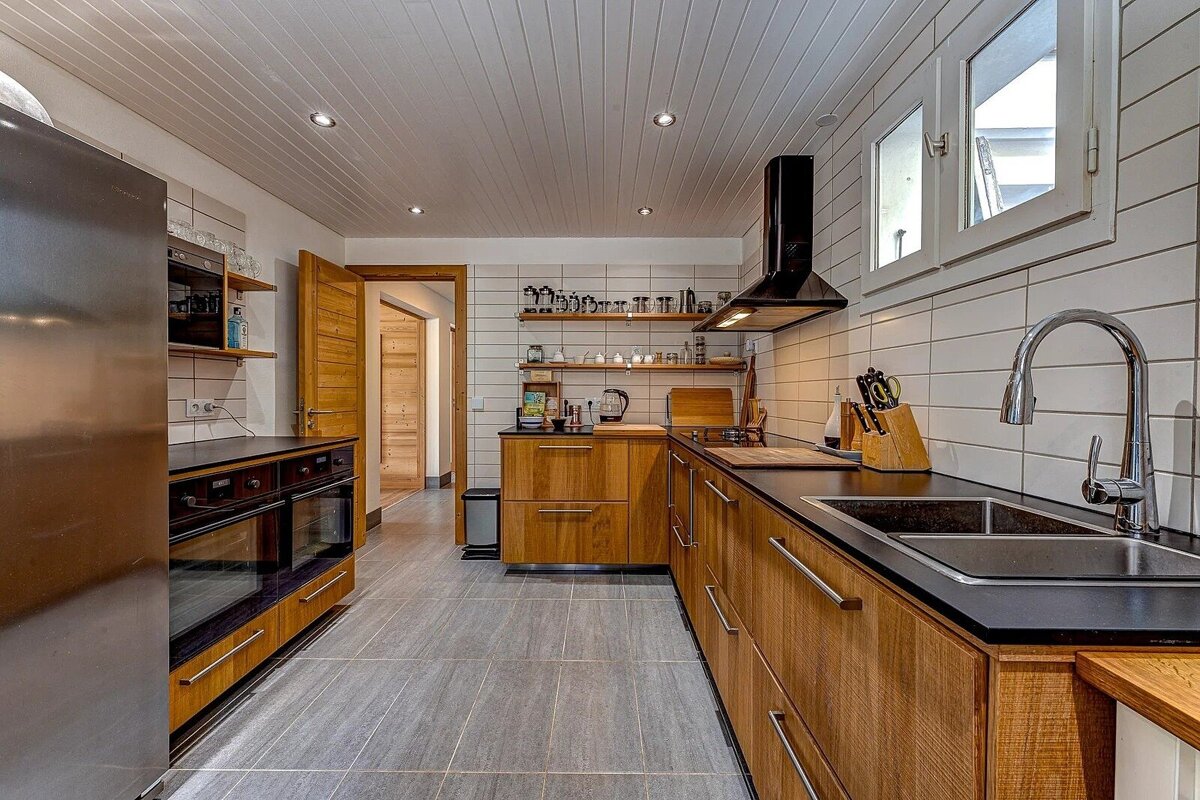 A modern kitchen with warm wooden cabinets, dark countertops, white tiled walls, and stainless steel appliances. Features a large sink and a window.