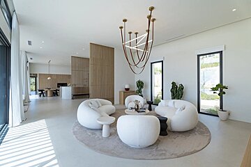 A bright, minimalist open-plan living room with white bouclé furniture, unique chandelier, and wood slat dividers. Dining area visible beyond.
