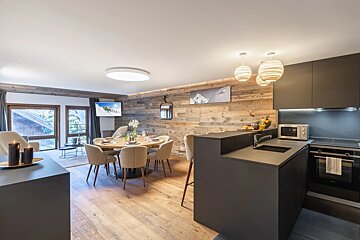 Chic open-plan chalet apartment with a modern dark kitchen, round dining table, and a living area featuring wood-paneled walls and large windows.
