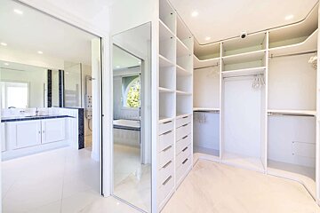 A clean, white walk-in closet with shelving and drawers, connecting to a bright modern bathroom with a vanity, shower, and reflected bathtub.
