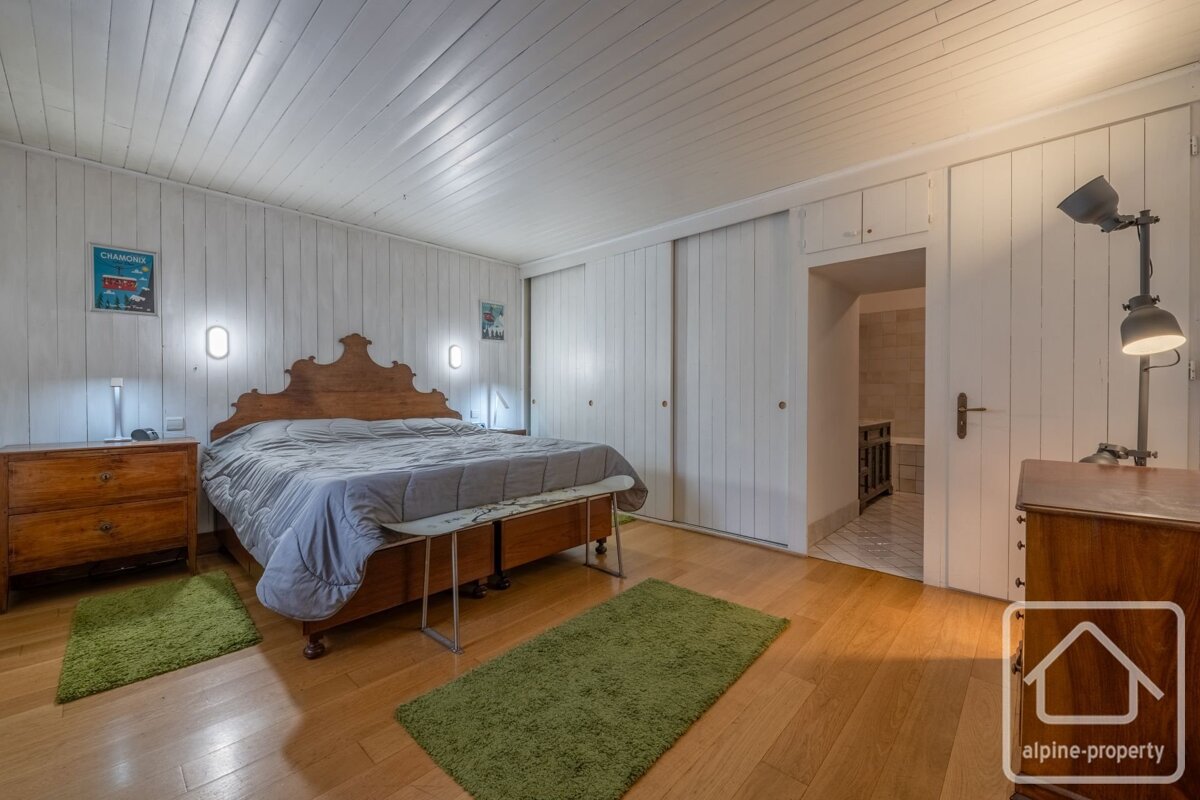 A cozy bedroom with white paneled walls and ceiling, a wooden bed, hardwood floors with green rugs, built-in wardrobes, and an open doorway.