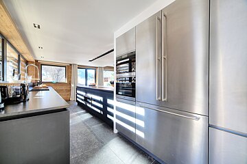 Bright, modern kitchen with stainless steel appliances, dark cabinetry, wood-paneled walls, and large windows casting sunlight on the tiled floor.