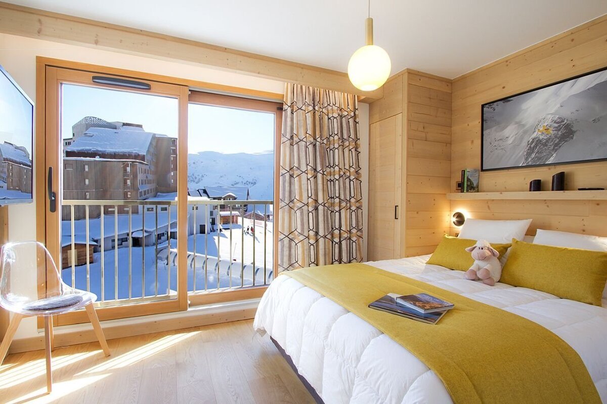 A warm, wood-paneled bedroom features a large window offering a stunning view of a snow-covered mountain resort, complemented by yellow accents and modern decor.