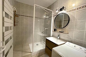 Tiled bathroom with a glass shower, sink vanity, washing machine, round mirror, and a wall-mounted towel warmer.