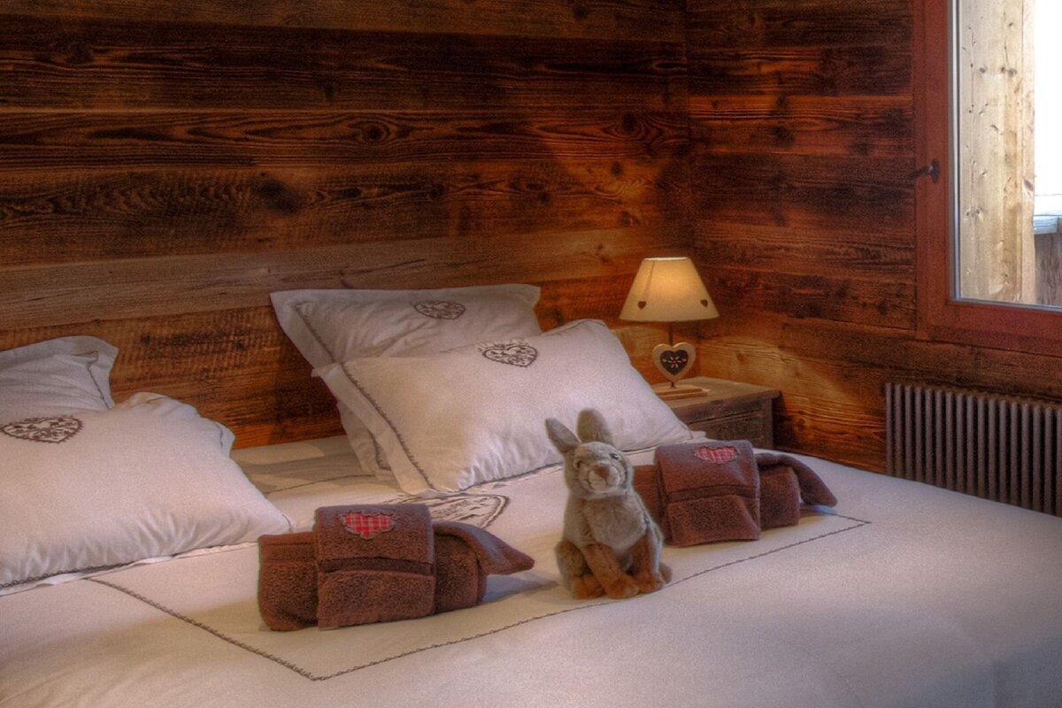 Cozy chalet bedroom featuring a bed with white linens, a stuffed bunny, folded towels, rustic wooden walls, and a warm lamp.