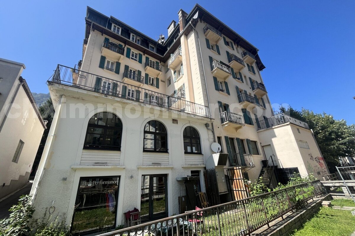 A tall, cream-colored building with multiple floors, green-shuttered windows, and balconies, featuring a ground-floor store, under a clear blue sky.