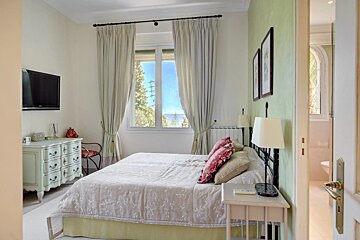 A light-colored bedroom with a bed, dresser, TV, and a window with curtains looking out to a sunny landscape.