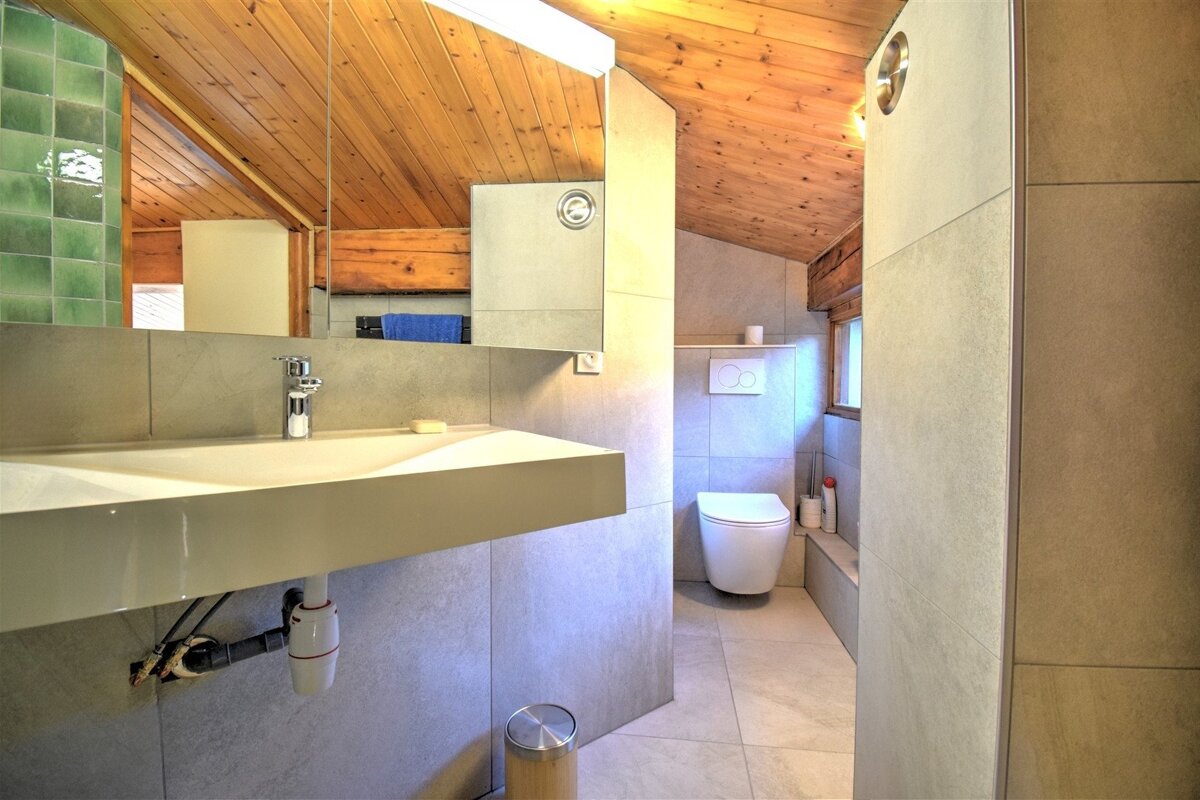 Modern bathroom featuring a floating sink, grey tile walls, a slanted wooden ceiling, and a toilet in the background.