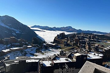 A mountain ski resort overlooks a valley filled with clouds, surrounded by snowy peaks under a clear blue sky.